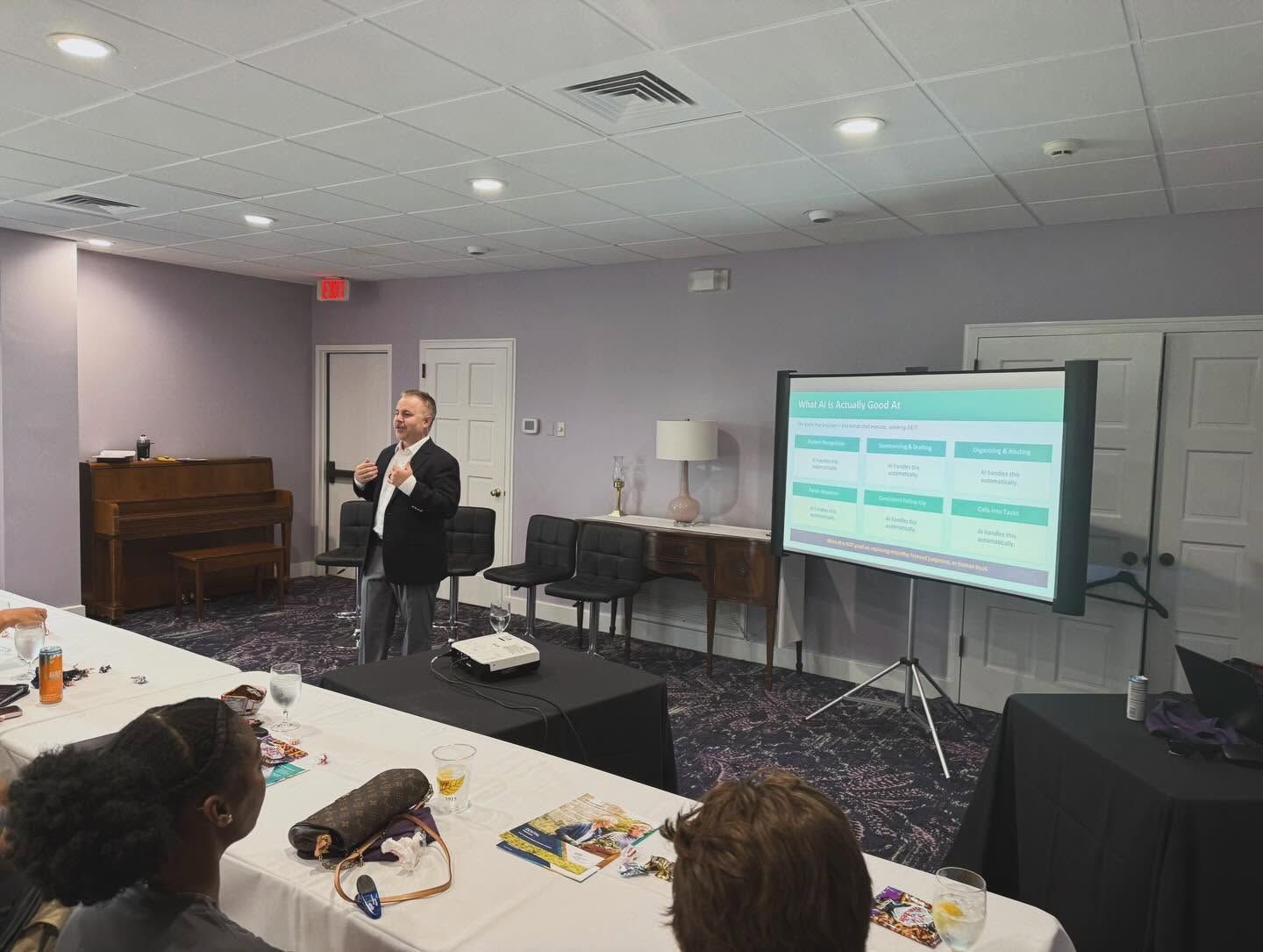 Jonathan Ferrell speaking at the front of a conference room next to a presentation screen, with attendees seated at a table in the foreground.